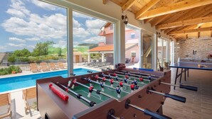 Une table de billard et une table de baby-foot se trouvent dans une pièce dotée de grandes fenêtres donnant sur une piscine et un paysage pittoresque.