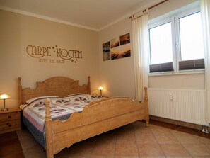 A bedroom with a wooden bed, a nightstand with a lamp, a window with curtains, and wall art. - Waldstr. Wa30-03 - Wa30-03 (Kühlungsborn)