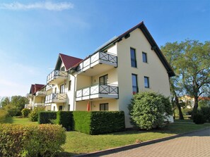 Um edifício residencial de vários andares com varandas, telhado de telhas vermelhas e um jardim bem cuidado.