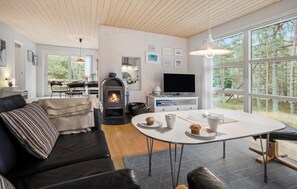 A modern living room with a fireplace, a dining area, and large windows offering a view of the forest. - Gorgeous home in Nexø with WiFi (Nexø)