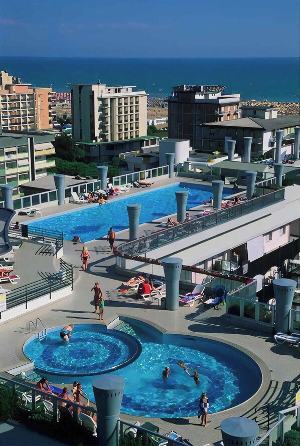A rooftop pool area with multiple swimming pools, lounge chairs, and a view of the ocean.