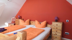 Un dormitorio con techo inclinado, dos camas con ropa de cama naranja, mesitas de noche de madera y una pared roja.