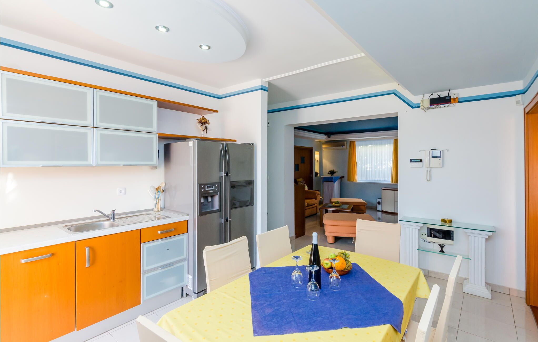 Une cuisine compacte dotée d’appareils électroménagers modernes, un coin repas avec une table préparée pour deux personnes, et un espace de vie avec un canapé et un téléviseur.