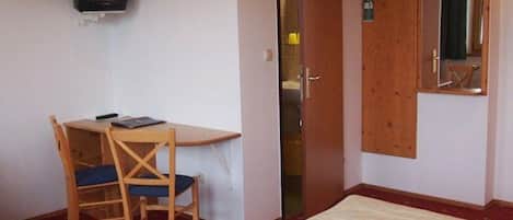 Una habitación con una cama, un escritorio con una silla, un televisor fijado a la pared y una puerta con un espejo.