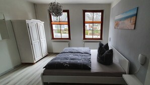 Een slaapkamer met een bed, twee ramen, een radiator, een witte inbouwkast en een schilderij aan de muur.