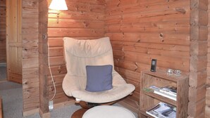 Una habitación acogedora tipo cabaña de troncos, con una butaca mullida, una mesita de noche con lámpara y una estantería de madera llena de libros.