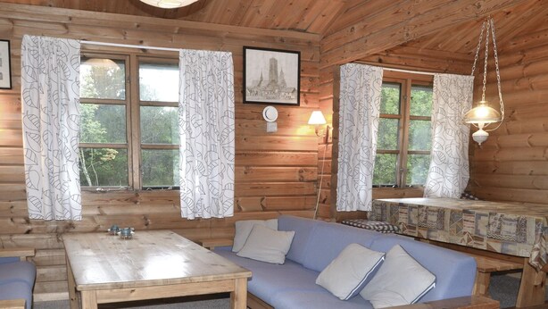Un interior de cabaña acogedor, con un sofá, una mesa y dos ventanas con cortinas estampadas de hojas.
