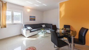 Una sala de estar moderna con un sofá gris, una mesa de centro de vidrio y sillas negras. Hay una ventana con cortinas, un cuadro en la pared y un suelo de baldosas.