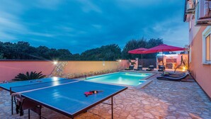 Una mesa de billar y una pequeña piscina en un espacio exterior con suelo de piedra, sillones de descanso y una parrilla.