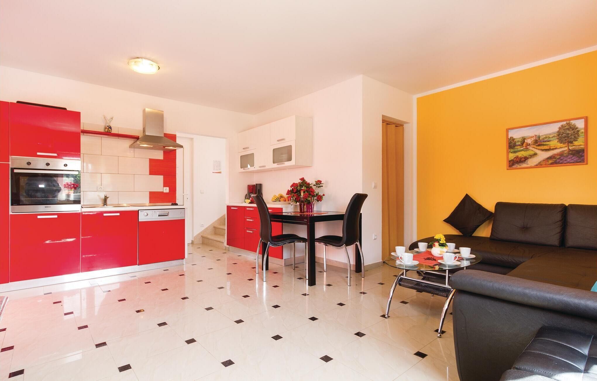 Een moderne keuken met rode kastjes, een eethoek met een zwarte tafel en stoelen, en een woongedeelte met een bank en een schilderij aan de muur.