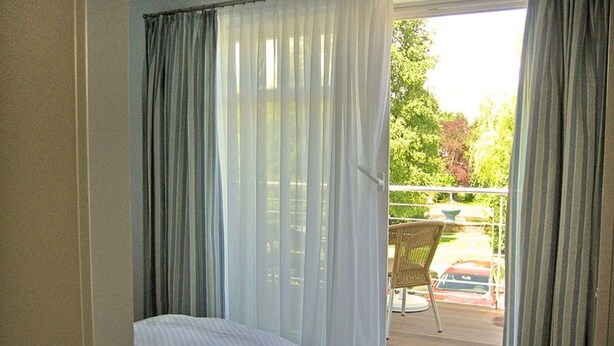 A room with a bed, a window with curtains, and a view of a balcony with a chair and a car. - Holiday Sundevit 1st - Holiday Sundevit (Prerow)