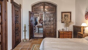 Une chambre à coucher avec un grand lit, une armoire en bois, un bureau avec une lampe et une porte donnant sur une autre pièce.