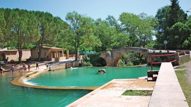 Una piscina con un ponte in pietra, circondata da alberi e un piccolo edificio.