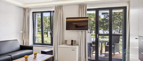 A modern hotel room with a sofa, a coffee table with flowers, a TV, a mini-fridge, and a sliding glass door leading to an outdoor seating area.