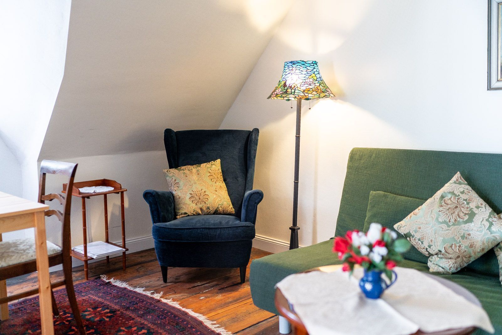 A cozy living room with a dark blue armchair, a green sofa, a wooden chair, and a floor lamp.