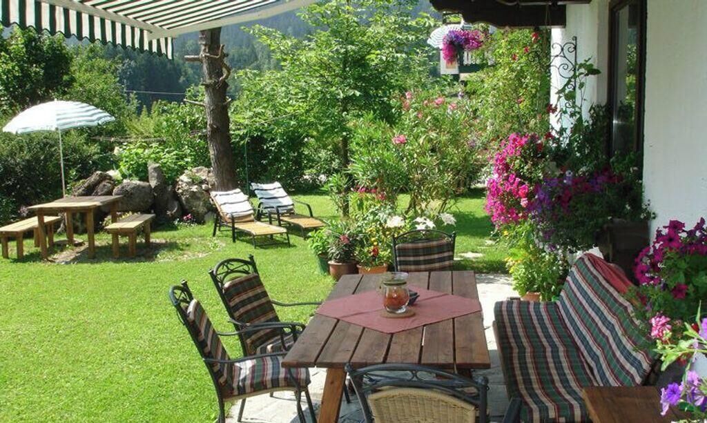 Eine Terrasse im Freien mit einem Holztisch und Stühlen, umgeben von üppigem Grün und blühenden Pflanzen.