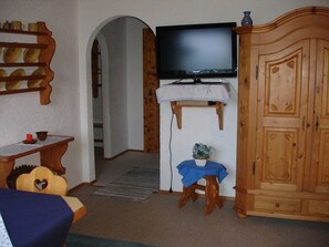 Ein Zimmer mit Fernseher auf einem Schrank, einem Holzkästen, einem kleinen Tisch mit blauer Tischdecke und einem Stuhl mit herzförmiger Rückenlehne.