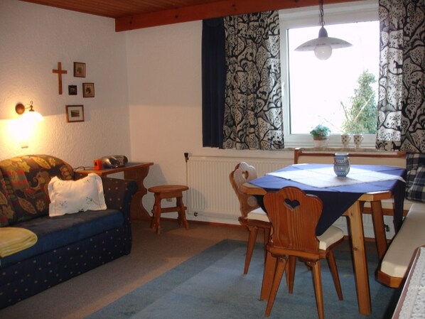 A cozy living room with a sofa, a dining table with chairs, a window with patterned curtains, and wall-mounted religious symbols.