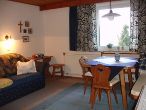 A cozy living room with a sofa, a dining table with chairs, a window with patterned curtains, and wall-mounted religious symbols.