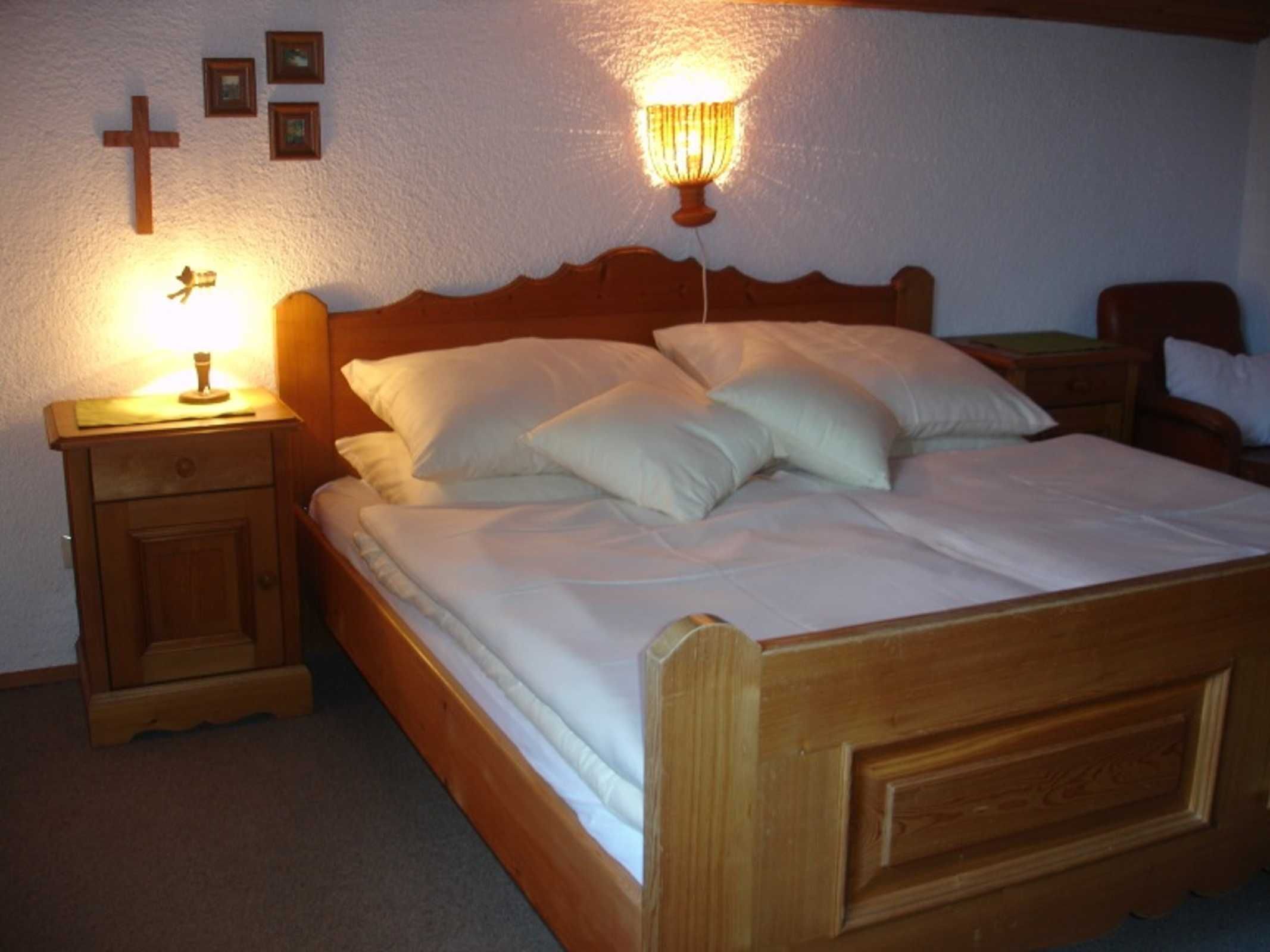 Ein hölzernes Bett mit weißer Bettwäsche, ein Nachttisch mit Lampe und ein an der Wand befestigtes Kreuz.
