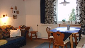 A cozy living room with a sofa, a dining table with chairs, a window with patterned curtains, and wall-mounted religious symbols. - Spatzennest apartment in country house style - Anna Laireiter (Keuschen)