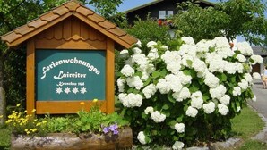 A wooden sign with text, a white hydrangea bush, and a house in the background. - Spatzennest apartment in country house style - Anna Laireiter (Keuschen)