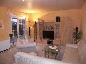 A living room with a glass coffee table, a sofa, a TV stand with shelves, and a sliding glass door leading outside.