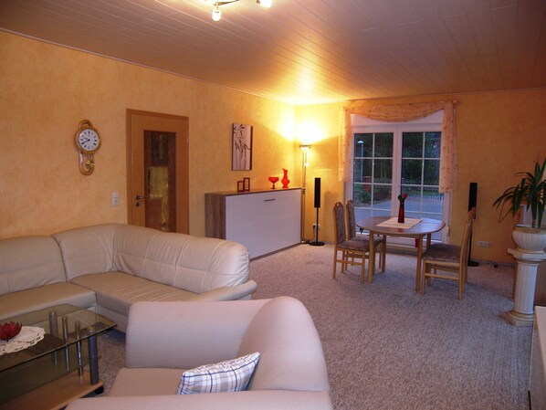 A living room with a sofa, a glass coffee table, a dining table with chairs, a lamp, and a clock on the wall.