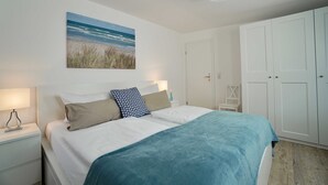 A neatly arranged bedroom with a bed, bedside lamps, a painting of a beach, and white wardrobes.