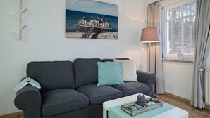 A living room with a grey sofa, a painting of a coastal house, a lamp, and a window with curtains.