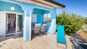 Ein blaues Gebäude mit einer Terrasse, auf der ein Tisch und Stühle stehen, einem Eingangsbereich und Blick auf Grünflächen.