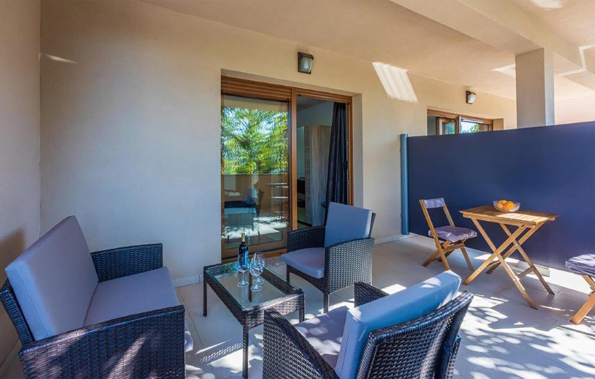 A sunlit outdoor seating area with wicker furniture, a glass table, and a view of greenery through a sliding glass door.