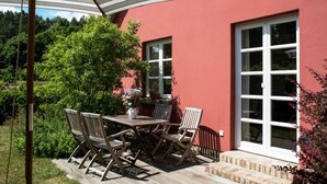 An outdoor patio with a wooden table and chairs, a striped umbrella, and a red building with large windows. - Landschek, Holiday home Das Ulmenhaus - Holiday home (Rankwitz)