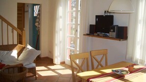 A cozy room with a wooden dining table, wicker chairs, a television on a shelf, and a window with curtains. - Landschek, Holiday home Das Ulmenhaus - Holiday home (Rankwitz)