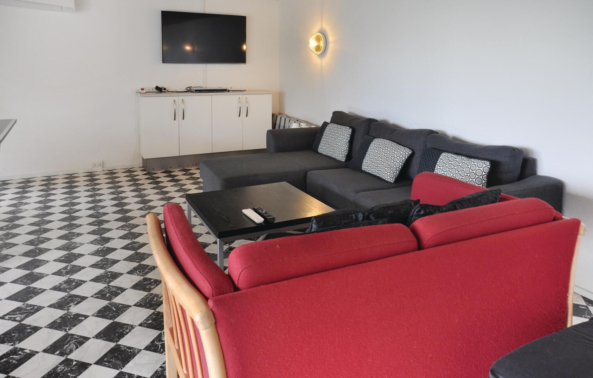 A living room with a checkered floor, a grey sofa with patterned cushions, a red armchair, a coffee table, and a flat-screen TV mounted on the wall.