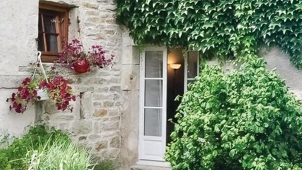 Een gebouw van steen met een witte deur en een raam met houten kader. Voor het gebouw hangt een plant met rode bloemen en staat een groene struik.