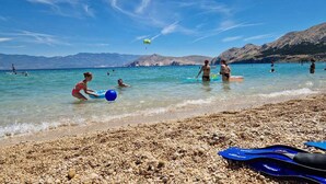 Une plage à l’eau d’un bleu limpide, avec un rivage de galets et des montagnes en arrière-plan.