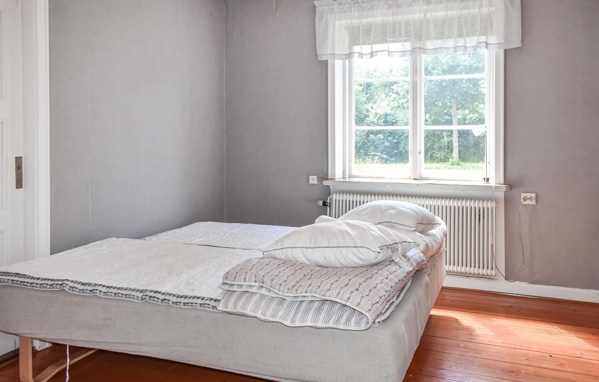 Una cama pequeña y bien hecha, con edredón y almohadas blancos, un radiador debajo de una ventana con cortinero blanco, y suelo de madera.