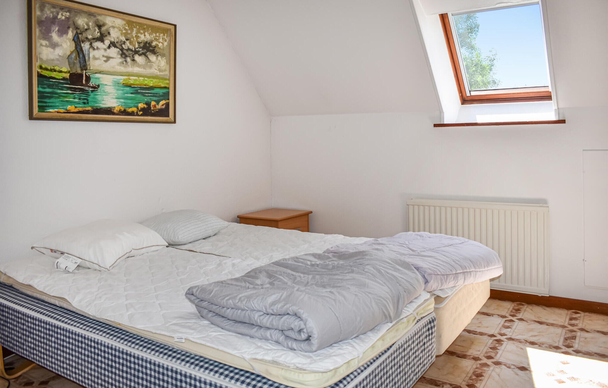 Un dormitorio con cama, mesita de noche, radiador, un cuadro y un tragaluz.