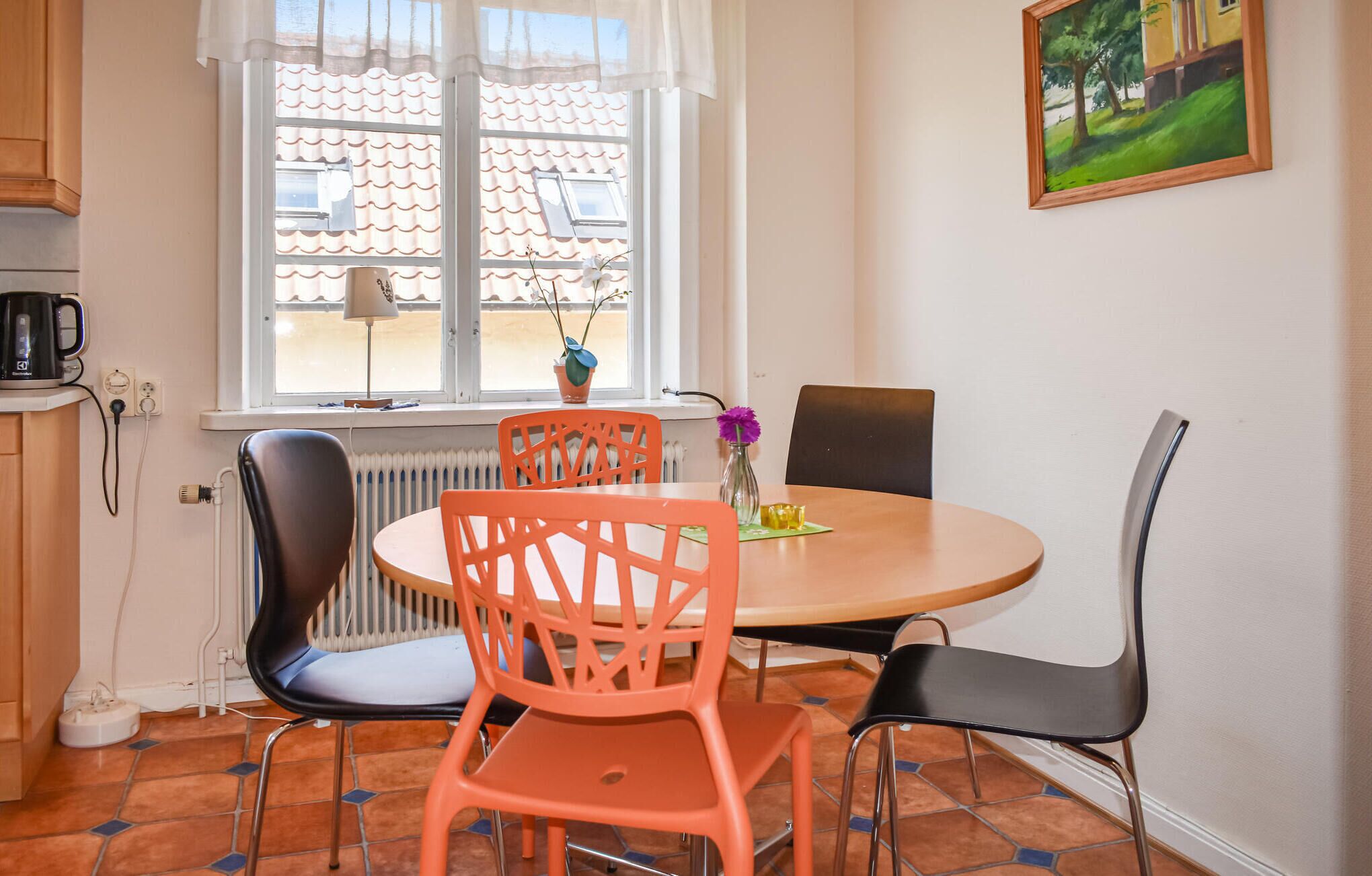 Un'area pranzo con un tavolo rotondo, sedie arancioni e nere, un termosifone, una finestra con vista su un edificio con tetto a tegole e un quadro appeso al muro.