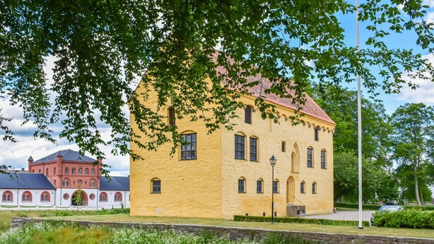 Un bâtiment jaune, doté de fenêtres en arc de cercle et d’un toit rouge, entouré de verdure et d’un mur de pierre.