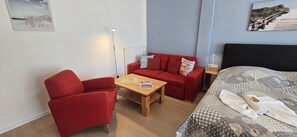 A room with a red sofa, a red armchair, a wooden table, a bed with a checkered duvet, and a floor lamp.