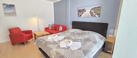 A hotel room with a bed, a red armchair, a small table, a red sofa, and two wall art pieces.