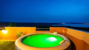 Ein Whirlpool im Freien mit grünem Innenraum, umgeben von einer Holzterrasse und einer gelben Wand, mit Blick auf ein Gewässer und entfernte Stadtlichter.