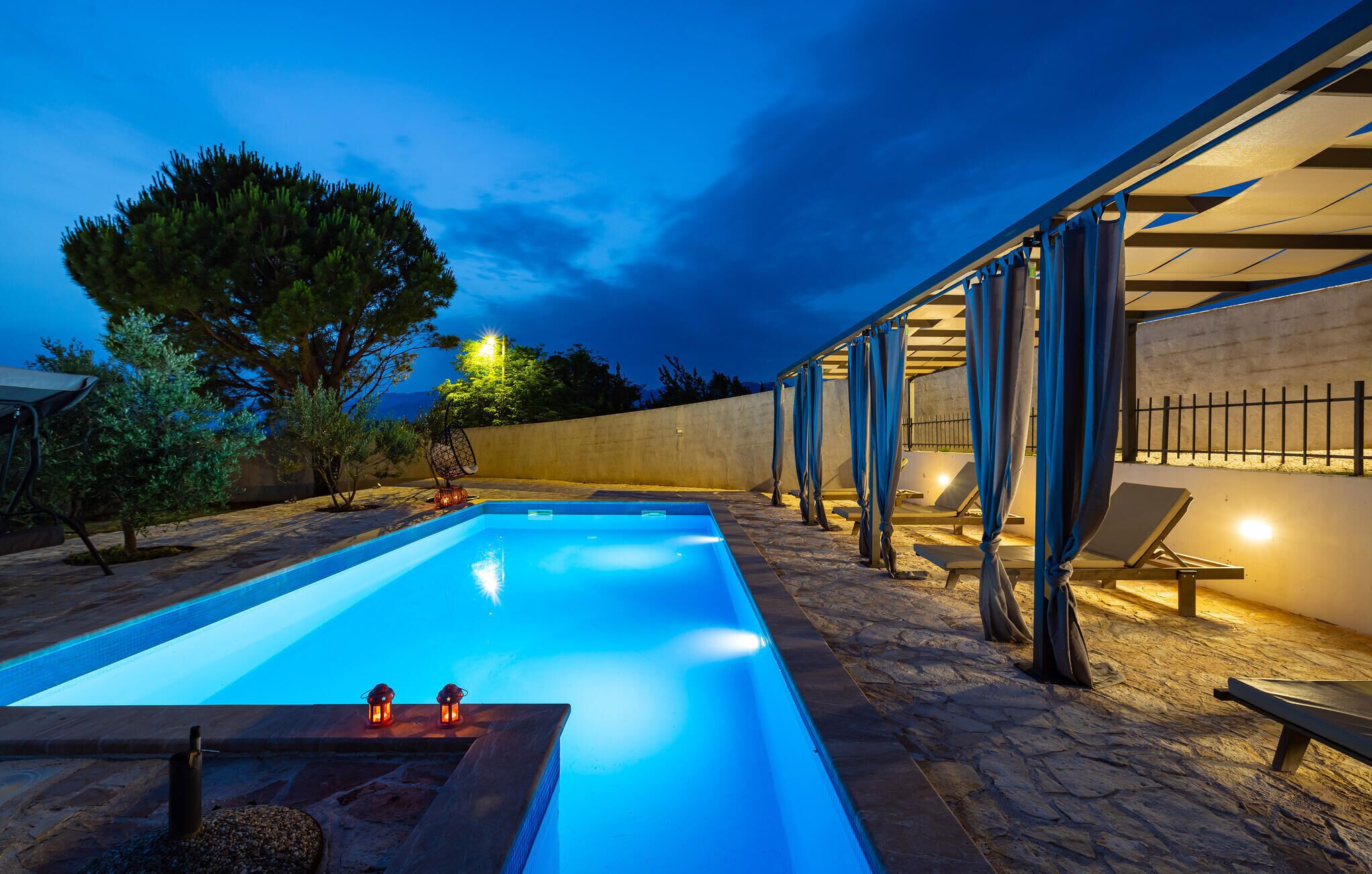 Ein rechteckiger Pool mit einer Steindeck, umgeben von Liegestühlen und einer Pergola mit blauen Vorhängen.