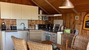 A wooden interior with a dining table, wicker chairs, a hanging light fixture, and kitchen appliances.