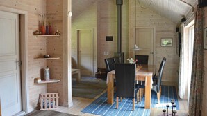 Habitació acollidora amb parets de fusta, taula de menjador amb caires, una petita estant amb objectes decoratius i finestra amb cortines.