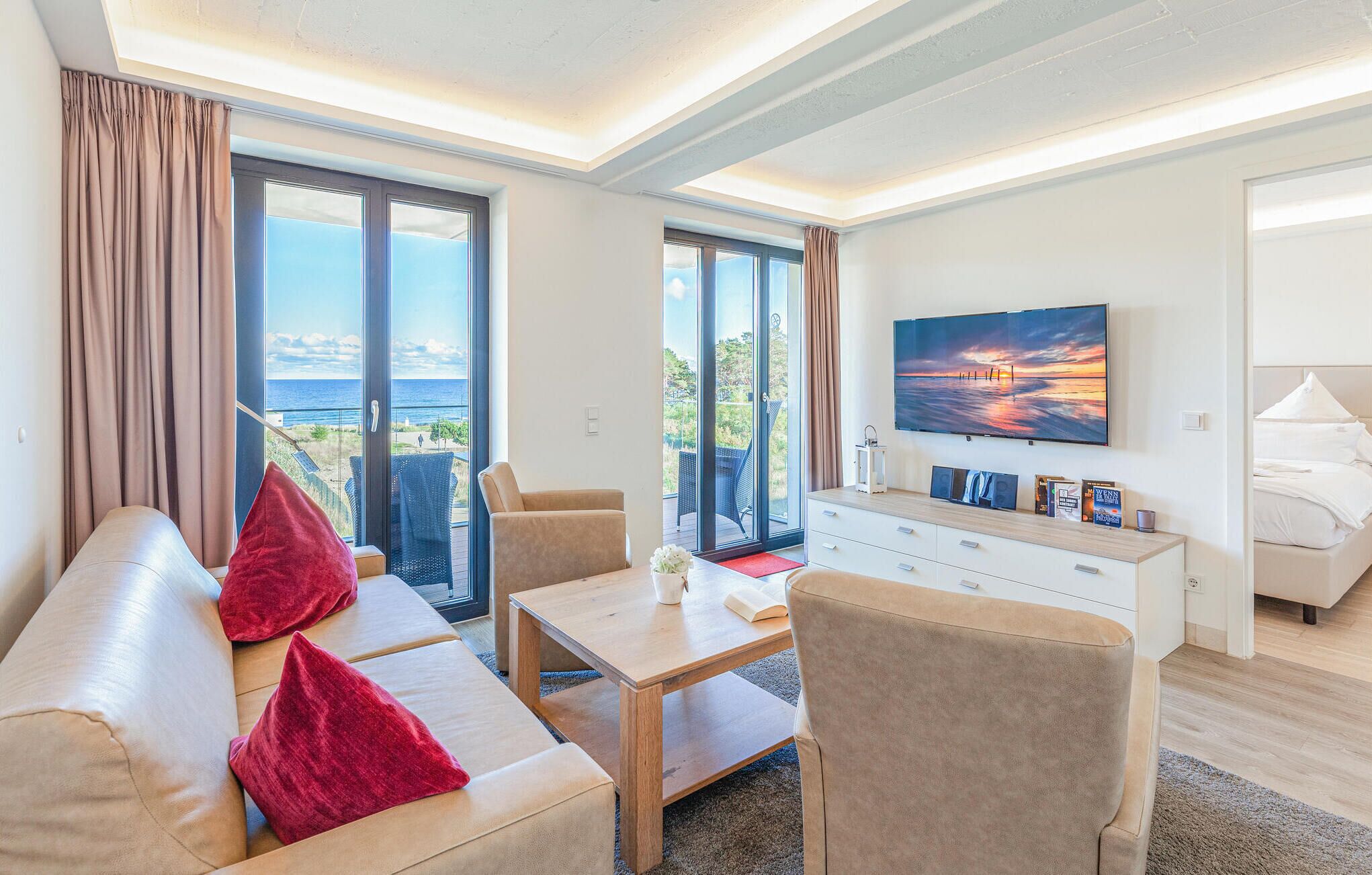 A modern living room with a sofa, armchairs, a coffee table, and a TV on a wall. There is a sliding glass door leading to a balcony with a view of the sea.