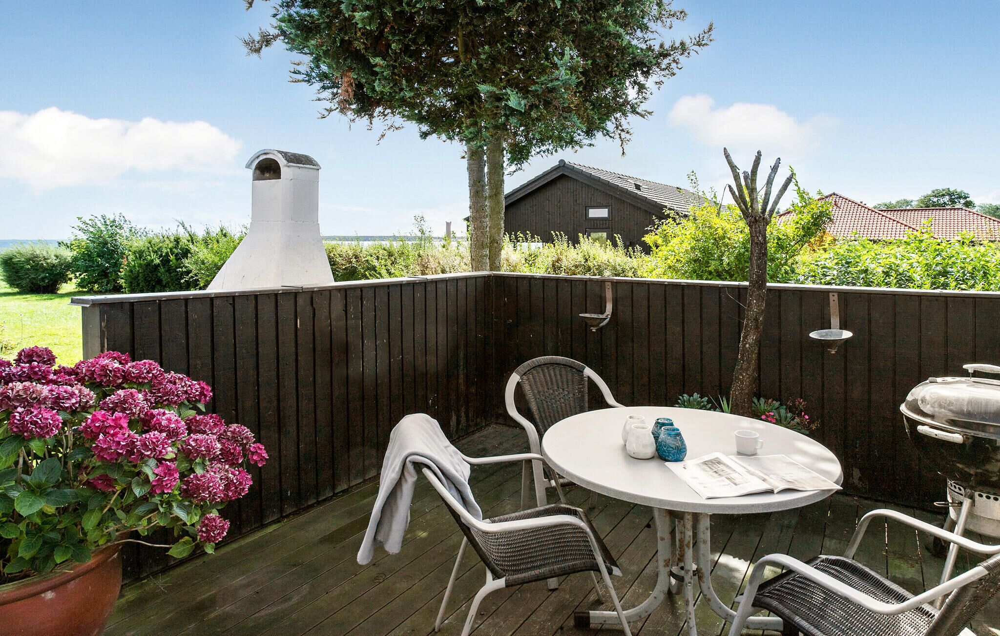En träaltan med ett runt bord och fyra stolar, en grill och en stor blomkruka med rosa blommor.