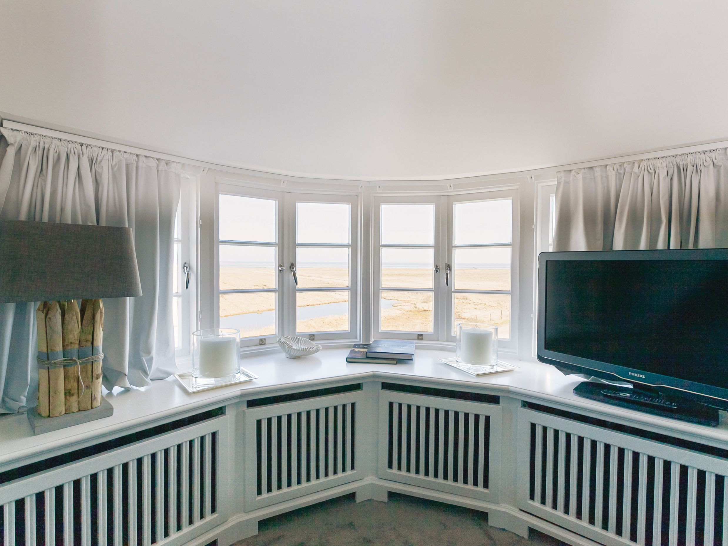 A corner room with a bay window overlooking a beach, a television, and a small table with a lamp and books.
